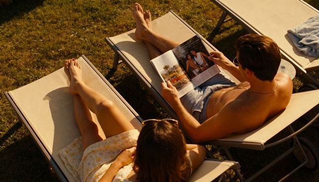 Sunlit couple in sundress and swim trunks lounging on lawn chairs reading magazine