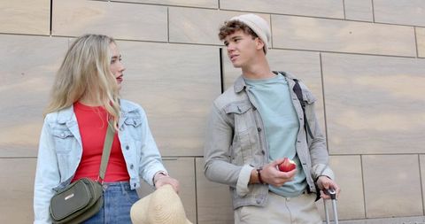 Young Couple Chatting on Urban Trip Leaning Against Stone Tile Wall Holding Luggage and Hat