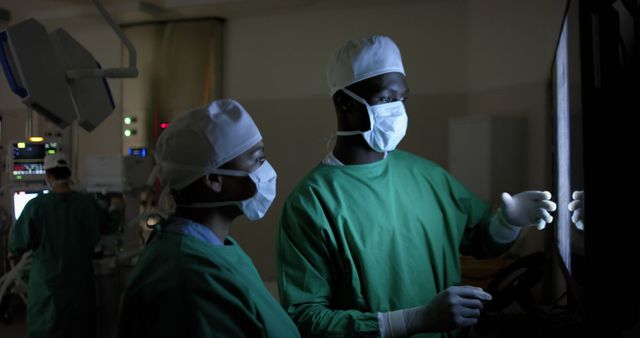 Two surgeons in green scrubs and masks are examining patient information on a monitor in an operating room. They appear to be reviewing crucial data, likely before commencing or during surgery. This can be used to depict themes of health care, medical technology, teamwork, and the critical environment of an operating room in various educational materials, medical journals, and hospital marketing content.