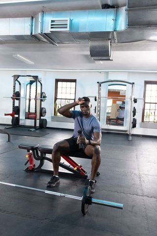 Fit african american man resting on bench holding shaker after barbell workout in modern gym
