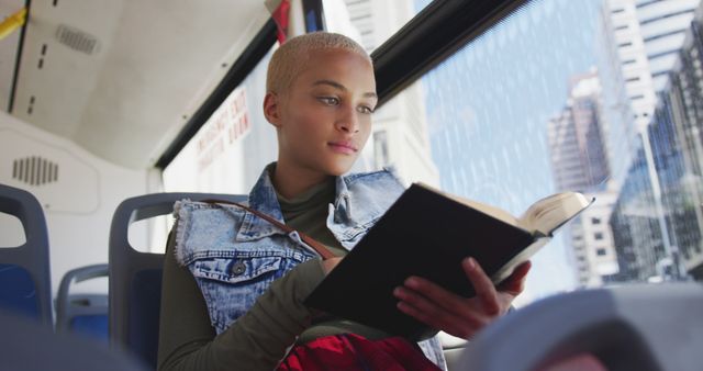 A woman with short blonde hair reads a book on a city bus during her commute. The bus has large windows showing tall buildings outside, capturing a busy urban setting. She is focused on her book, wearing a denim jacket over a green top. This image is perfect for illustrating public transportation, urban commuting, reading habits, and lifestyle themes.