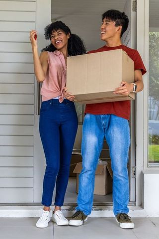 Young couple unlocking new home while carrying moving box on porch, celebrating move-in