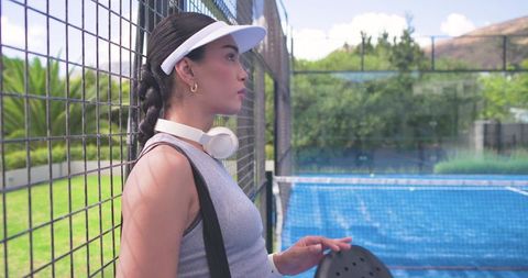 Young female paddle tennis player leaning against court fence wearing visor and headphones