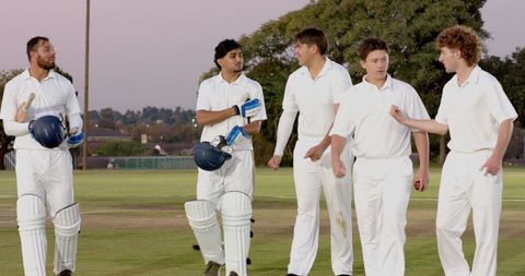 Cricket team walking across pitch at dusk in white uniforms carrying helmets and bat