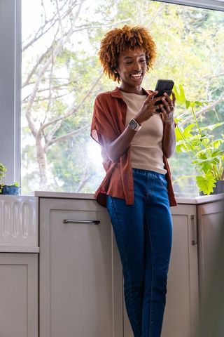 Sunlit african american woman leaning on kitchen counter, texting on smartphone, smiling