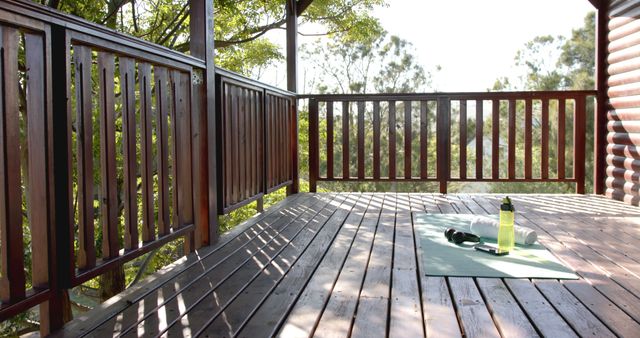 Outdoor yoga space set on a wooden deck with a water bottle and a yoga mat. Trees and sunlight filter through slatted wooden railings, creating a serene atmosphere. Perfect for visuals focused on fitness, wellness retreats, peaceful living, or outdoor exercise tutorials.