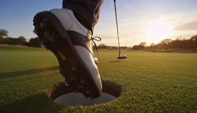 Golfer stepping over cup with spiked shoe on putting green at sunset