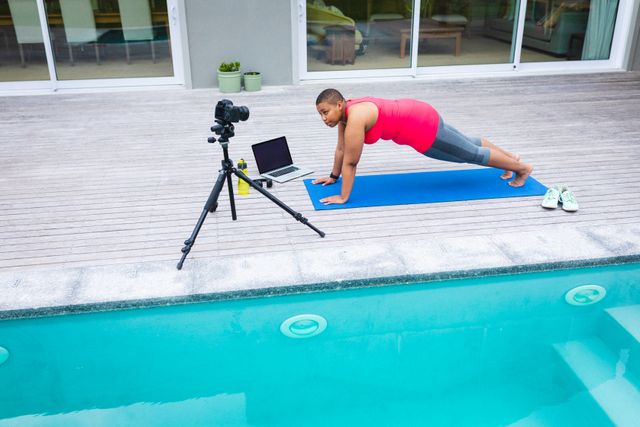 African American woman recording a fitness video by the poolside, demonstrating exercises on a yoga mat. Ideal for use in content related to fitness, healthy lifestyle, home workouts, vlogging, and influencer marketing.