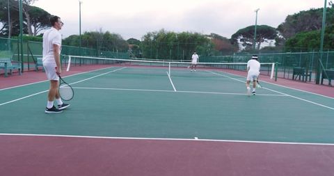 Diverse male tennis players playing doubles on outdoor hard court in white uniforms