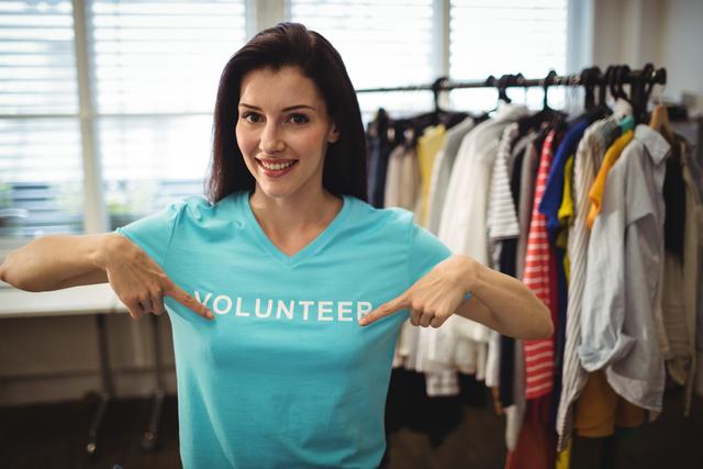 Female volunteer proudly pointing at her 'Volunteer' t-shirt in a clothing donation center. Ideal for use in materials promoting volunteerism, charity events, community service initiatives, and nonprofit organizations. Perfect for illustrating themes of giving back, social responsibility, and humanitarian efforts.