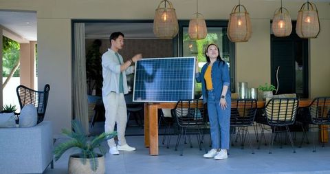 Asian couple inspecting freestanding solar panel on modern covered patio with dining area