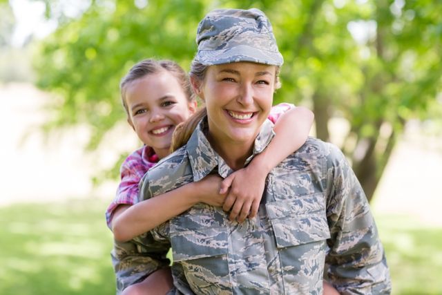 Female soldier in camouflage uniform giving a piggyback ride to her smiling daughter in a park. Perfect for themes of military families, parent-child bonding, outdoor activities, and family happiness. Ideal for use in articles, advertisements, and social media posts about military life, family support, and summer fun.