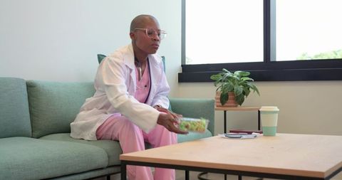 African american nurse taking lunch break on clinic sofa reaching for salad container