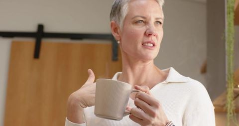 Mature woman holding ceramic mug and gesturing in cozy minimalist interior morning light