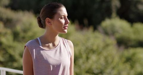 Young woman squinting toward sun on balcony wearing bun and pink sleeveless top
