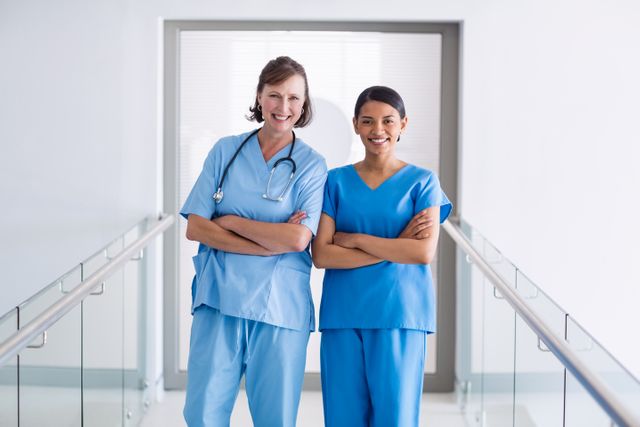 Two healthcare professionals, a nurse and a doctor, are standing in a hospital corridor with their arms crossed, smiling at the camera. Both are wearing blue scrubs, indicating a professional medical environment. This image can be used for promoting healthcare services, medical team collaboration, hospital advertisements, or educational materials related to healthcare professions.