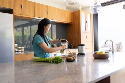 Mature african woman preparing green smoothie in modern kitchen with marble countertop