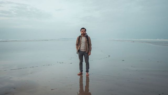 Solitary traveler standing barefoot on misty coastal sand with backpack and reflection