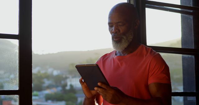Mature man in casual red t-shirt standing by window while using digital tablet with serene backdrop of sunset and subtle cityscape. The gentle backlight highlights concentration and tranquility. Perfect for promoting tech products, modern lifestyle, relaxed environments, and age-focused wellness concepts.