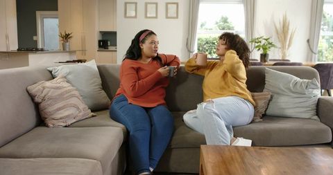Diverse female friends relaxing and chatting on gray sectional sofa while holding mugs