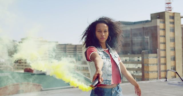 Woman confidently holding yellow smoke bomb on rooftop with city buildings in background. Ideal for urban lifestyle, street fashion, self-expression, empowerment, and city culture themes.