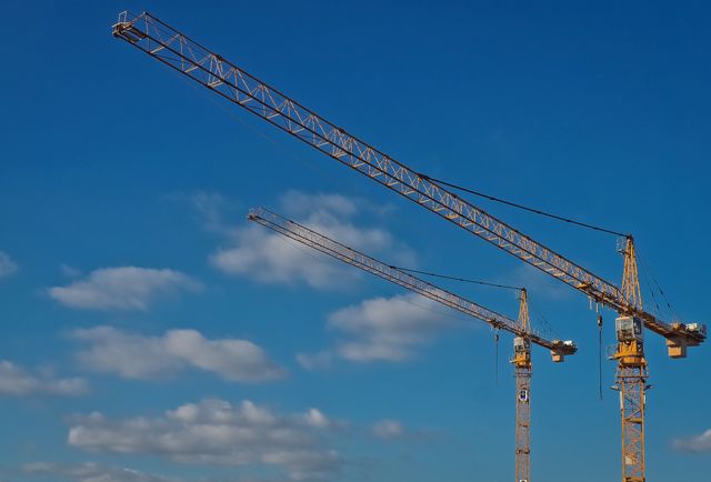 Tower cranes extending into clear blue sky, emphasizing construction and development. Useful for illustrating themes related to industrial equipment, building projects, engineering, and infrastructure development. Ideal for use in publications or content about construction industry, technology in construction, and urban development.