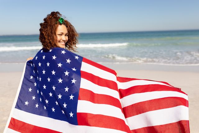 Biracial woman with curly brown hair and light brown skin holding an American flag on a beach, looking back with a smile. She is wearing a green hair accessory. Perfect for themes of patriotism, summer celebrations, beach vacations, and American holidays like Independence Day and Memorial Day.