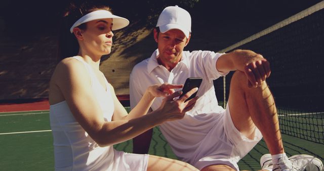 A Caucasian man and woman, both dressed in tennis attire, are sitting on a tennis court reviewing something on a smartphone, with copy space. Their focused expressions suggest they could be analyzing their performance or planning strategies.