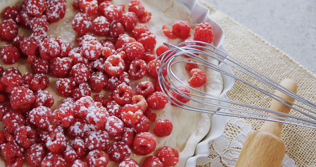 A raspberry tart is being prepared, with fresh raspberries sprinkled with powdered sugar next to a whisk and a rolling pin, with copy space. Culinary enthusiasts and home bakers often enjoy creating such desserts, which combine the tartness of fruit with the sweetness of sugar.
