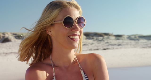 A young Caucasian woman enjoys a sunny day at the beach, wearing stylish sunglasses and a smile, with copy space. Her carefree expression and the coastal backdrop evoke a sense of relaxation and summer vibes.