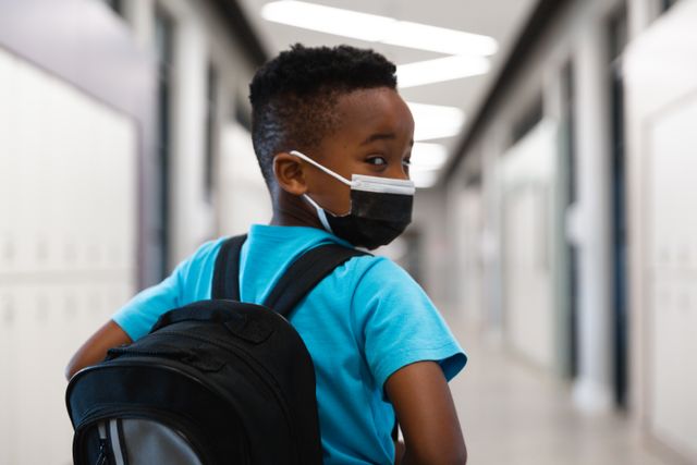 African American schoolboy wearing a mask and carrying a backpack while looking over his shoulder in a school hallway. This image can be used for educational materials, health and safety campaigns, back-to-school promotions, and articles related to the impact of COVID-19 on education and childhood.
