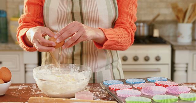 Woman cracking eggs into mixing bowl as she bakes cupcakes in home kitchen. Baking tray with colorful cupcake liners placed on counter. Ideal for blogs and articles on homemaking, baking tutorials, recipe websites, and domestic life representations.