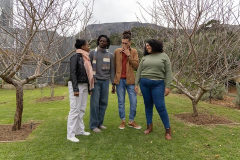 Diverse friends chatting and laughing on orchard lawn wearing jackets and scarves