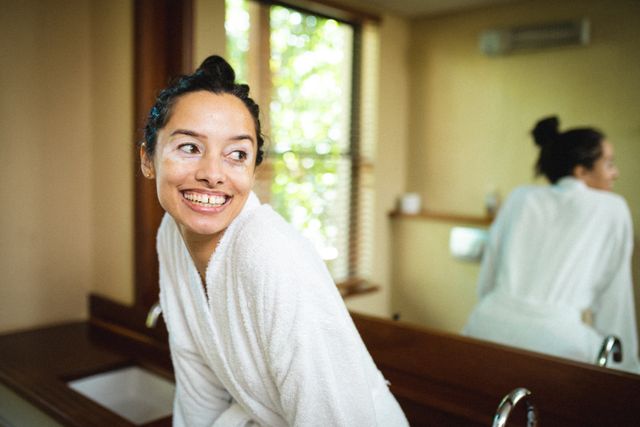 Young biracial woman with vitiligo smiling while sitting on bathroom vanity in a bathrobe. Ideal for promoting body positivity, self-care routines, wellness products, and natural beauty. Perfect for use in lifestyle blogs, spa advertisements, and skincare campaigns.