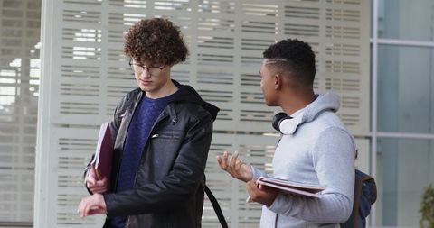 Two College Students Conversing Outside Modern Campus Holding Notebooks and Headphones