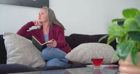 Mature woman relaxing on couch holding notebook and drinking tea in cozy living room