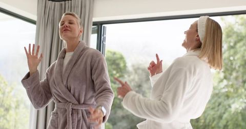 Mature friends laughing and dancing in bathrobes by sunlit floor-to-ceiling glass doors