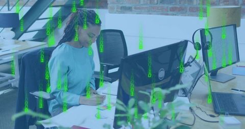 Woman taking notes at modern office desk with dual monitors, laptop and green data overlay