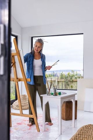 Mature woman painting at easel in light-filled studio, holding brushes, evaluating canvas