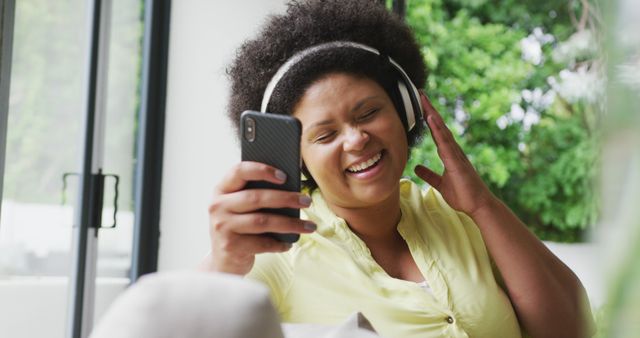 Image of happy plus size african american woman with headphones on sofa, listening to music. lifestyle, leisure, spending free time at home with technology.