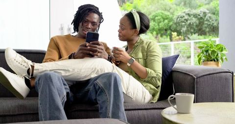African American Couple Relaxing on Sofa Sharing Smartphone and Coffee on Balcony