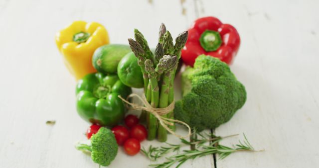 Arrangement of vibrant fresh organic vegetables including yellow, green, and red bell peppers, asparagus, broccoli, cherry tomatoes, lime, and rosemary, on rustic white wooden background. Ideal for promoting healthy eating, vegetarian and vegan lifestyles, organic farms, grocery stores, and food-related blogs or websites.