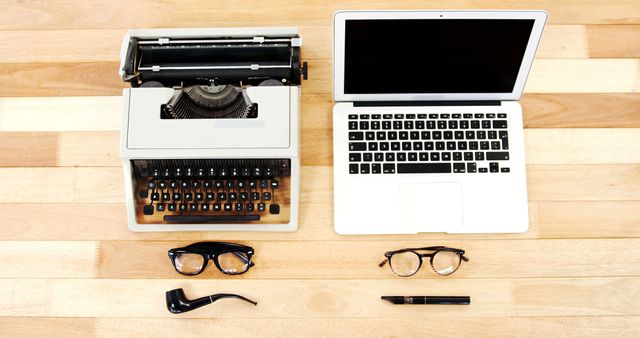 This stock image features a top view of a vintage typewriter paired with a modern laptop on a wooden desk. Two pairs of glasses, a pipe, and a fountain pen are also neatly placed on the desk. This image is ideal for illustrating the evolution of technology, the contrast between old and new, and themes of communication and writing. It is perfect for blogs, articles, or marketing materials related to technology, retro styles, and office workspace aesthetics.