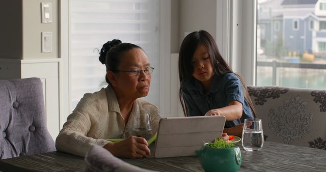 Front view of cute asian granddaughter and old grandmother using digital tablet while eating food. Food plate and glass of water on dining table 4k