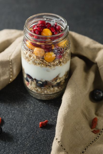 Layered yogurt parfait with pomegranates, golden berries, and granola in a mason jar. Ideal for promoting healthy breakfast options, nutritious snacks, or homemade desserts. Perfect for food blogs, recipe websites, and health-focused publications.