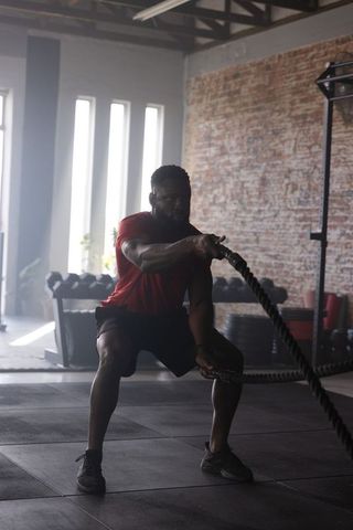 Muscular athlete performing battle rope waves in industrial gym loft with dumbbell rack