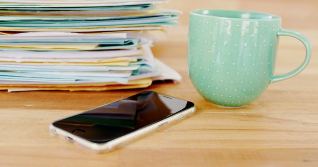 A stack of documents sits next to a smartphone and a mint green coffee mug on a wooden table, with copy space. This setup suggests a work environment where someone might be taking a break or multitasking between paperwork and digital communication.