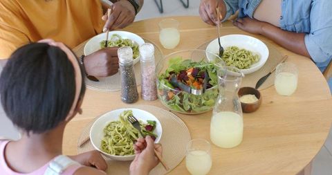 African family enjoying pesto pasta and salad around round wooden dining table