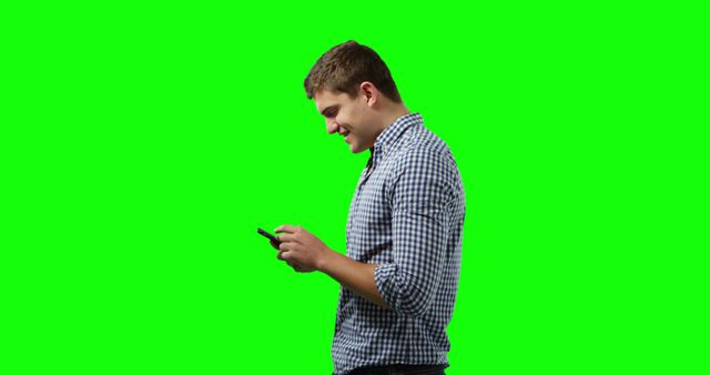 Young man in casual checkered shirt using smartphone, standing against a green screen. Ideal for technology, communication, social media, and user interface design content. Green screen background can be easily replaced with any desired imagery or video.