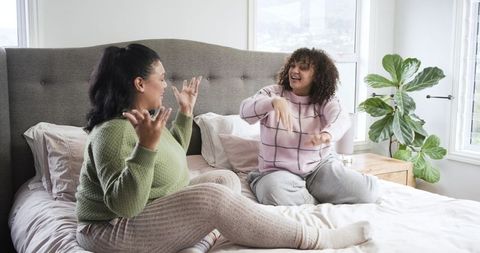 Two african american women laughing and chatting on cozy bed in sunlit bedroom